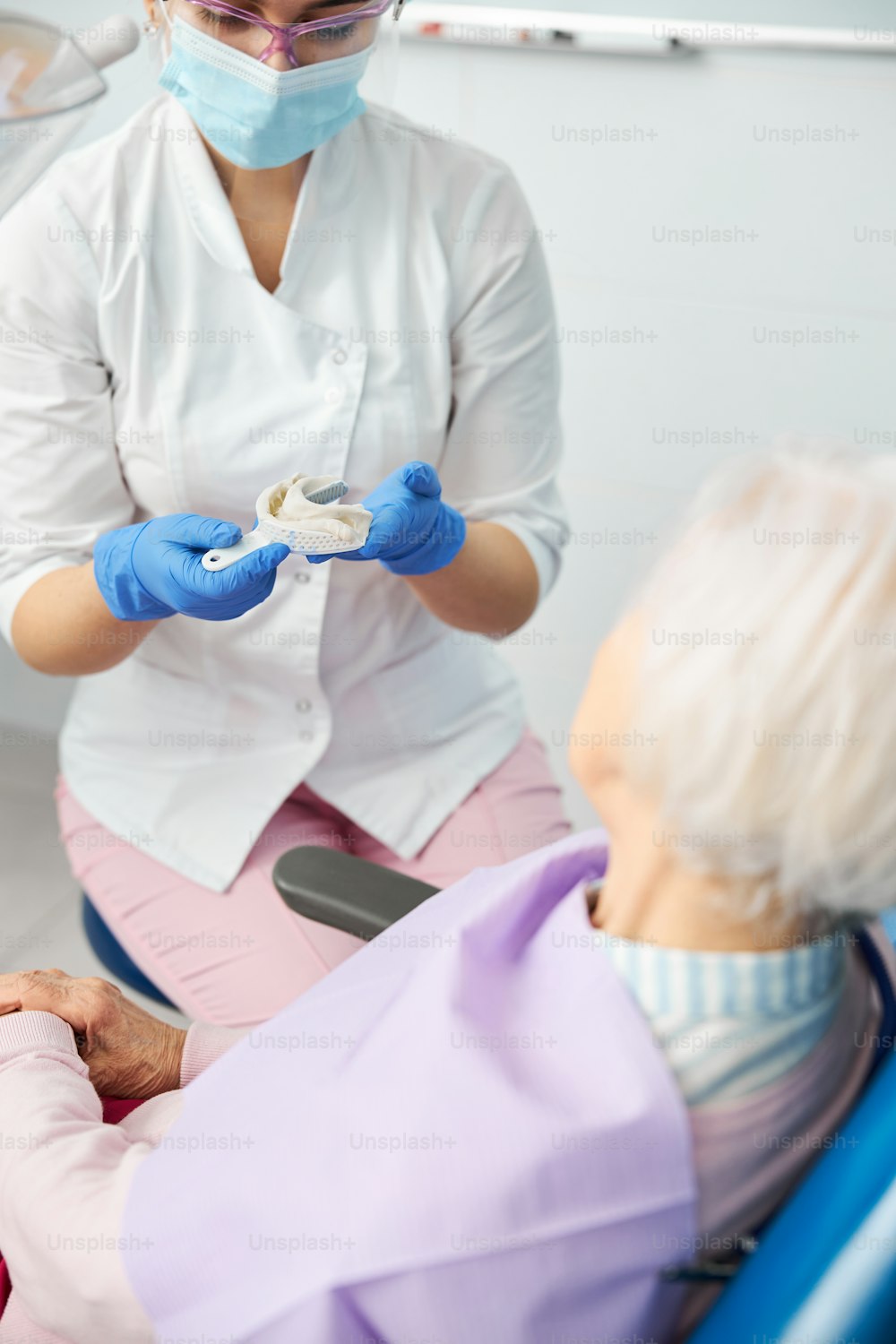Medico dentale che si siede su una sedia accanto a un paziente anziano e studia l'impronta in silicone di una mascella