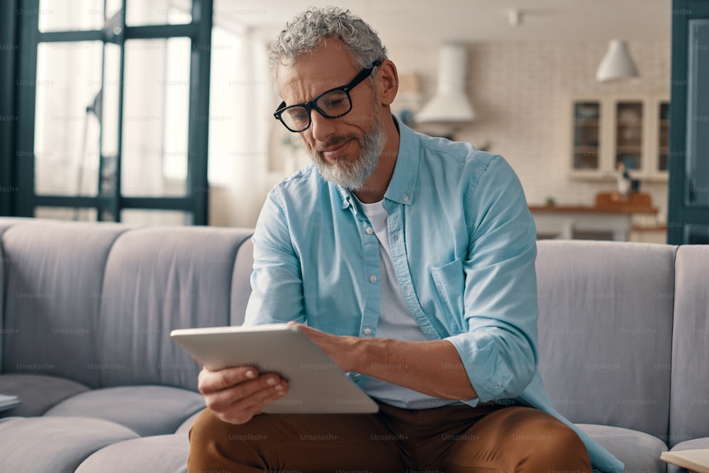 Hombre mayor concentrado en ropa casual usando tableta digital mientras está sentado en el sofá de casa