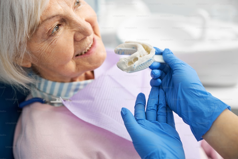 Empleada médica señalando con la palma de su mano la huella de un diente que sostiene cerca de la barbilla de la jubilada
