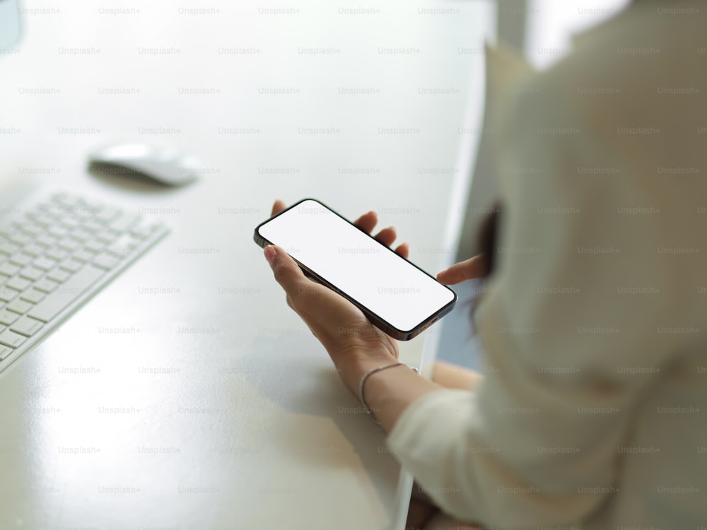 Cropped shot of female hand holding smartphone with mock up screen on computer desk, clipping path