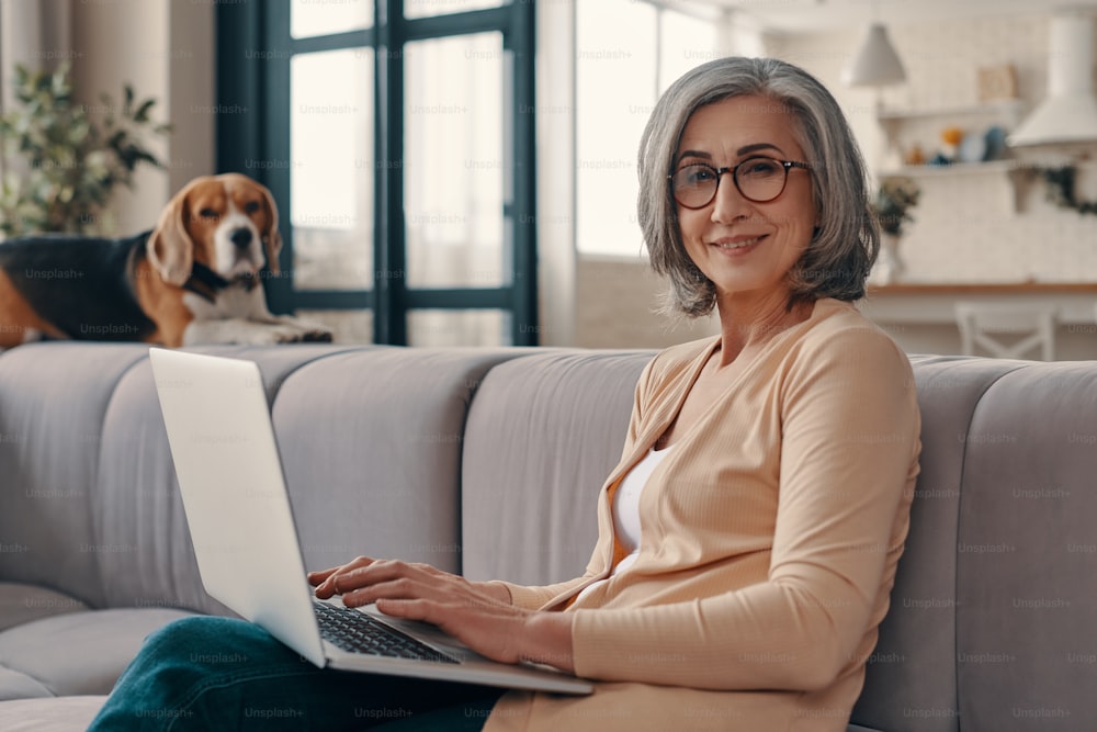 Cheerful senior woman in casual clothing using laptop and looking at camera while sitting on the sofa at home