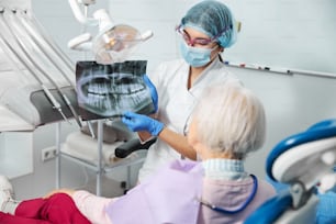 Dama en traje médico con protector facial que mantiene el diagrama de rayos X de los dientes antes de la cara de la persona mayor mientras describe su condición