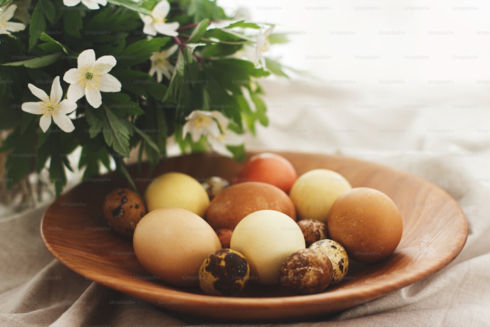 Modern easter eggs in wooden bowl on rustic linen cloth with spring flowers. Happy Easter! Natural dyed eggs in yellow and red colors on rural table with blooming flowers anemone. Aesthetic