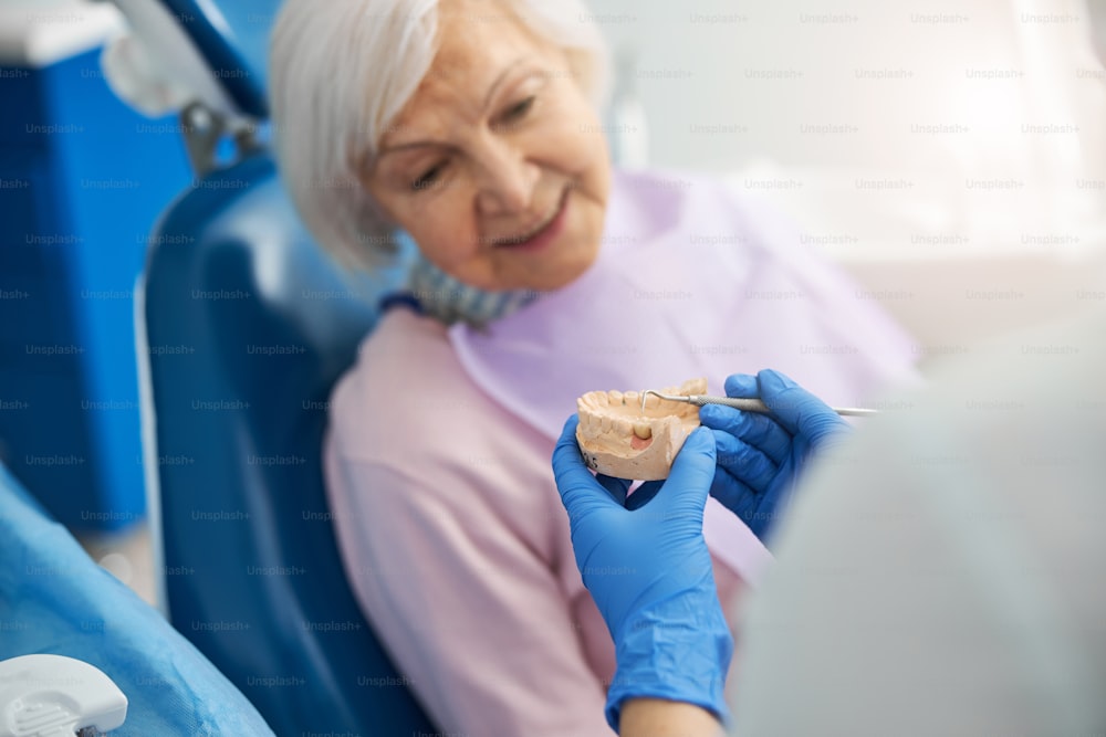 Cirujano dental que usa una sonda periodontal para apuntar un diente enfermo en un molde de mandíbula inferior para una dama mayor