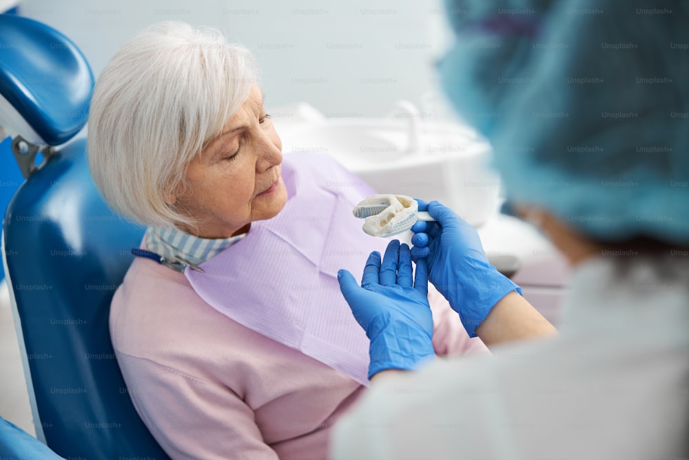 Dental surgeon offering elastic tooth impression from silicone to a retired woman in operation chair