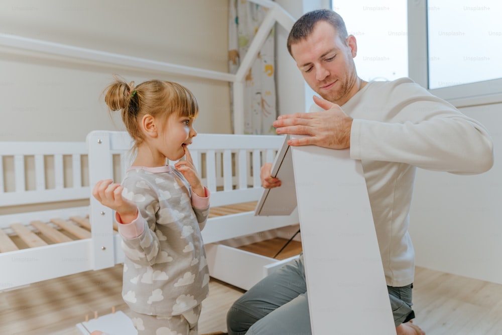 小さな4歳の女の子は、父親が子供部屋のベッドの引き出しを組み立てたり修理したりするのを手伝います。