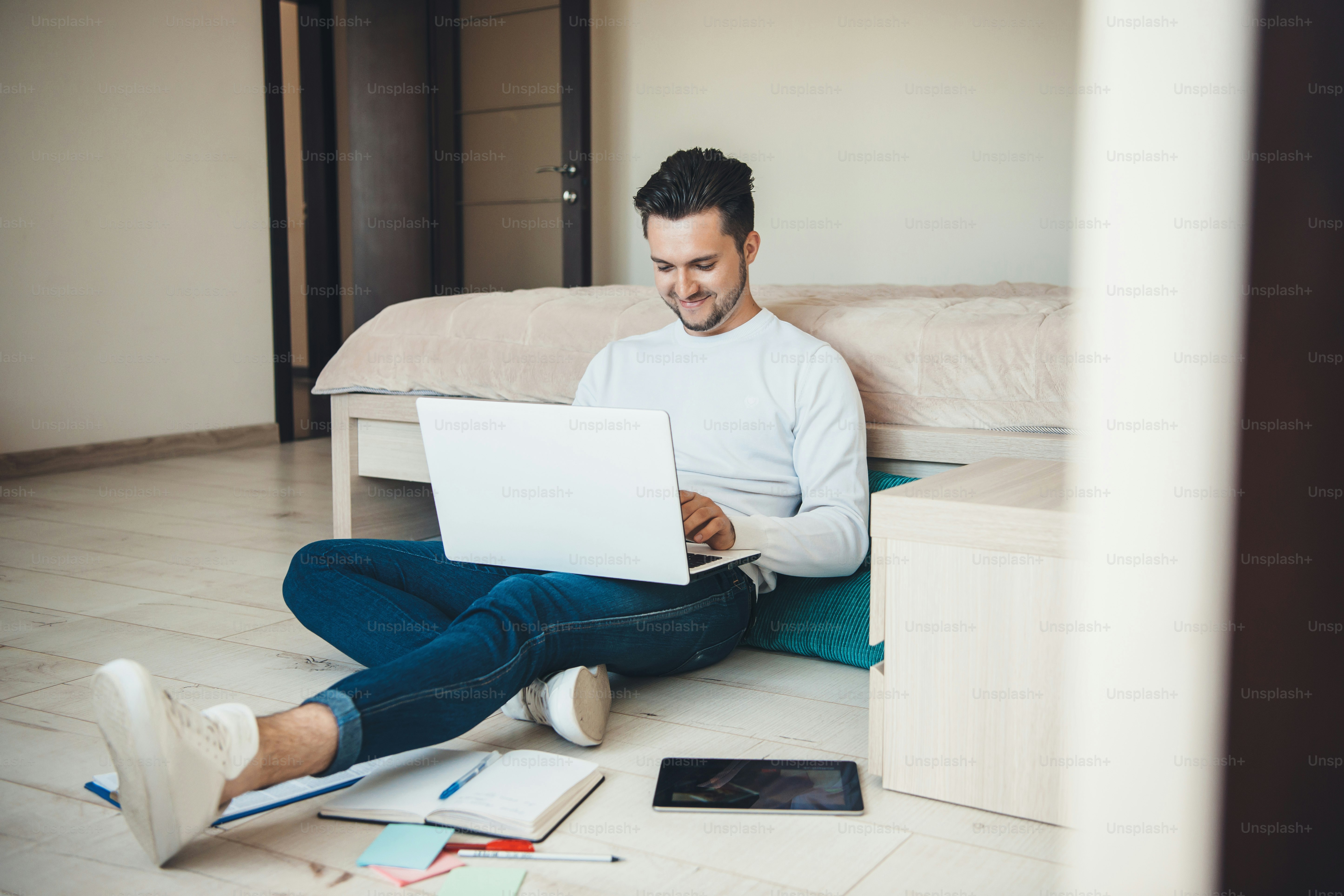 Smiling caucasian man is sitting on the floor working with a laptop remotely and some papers