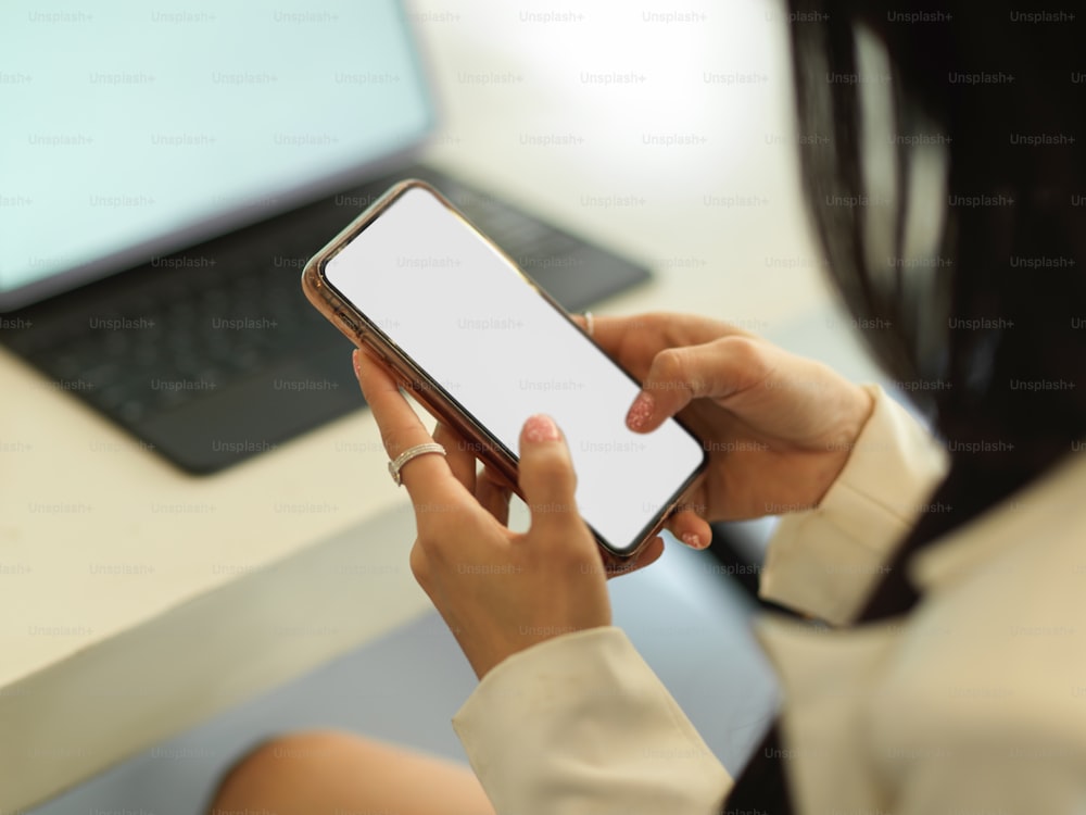 Abgeschnittene Aufnahme von Geschäftsfrauenhänden, die SMS auf einem Mock-up-Smartphone schreiben, während sie am Arbeitsplatz sitzen, Pfad abschneiden