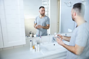 Hübsche männliche Person, die eine Flasche Duft hält, während sie am Waschbecken steht und einen lustigen Ausdruck macht