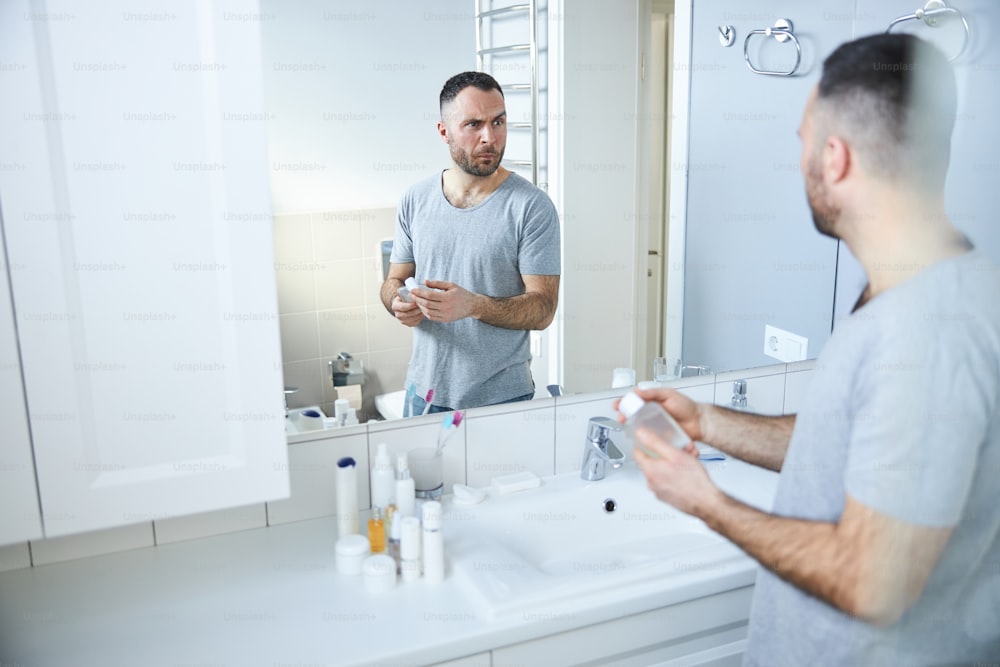 Handsome male person holding bottle of fragrance while standing by the sink and making funny expression