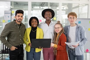 Portrait of happy multiethnic creative people with documents and modern laptop standing at office, smiling and looking at camera. Concept of technology and cooperation.