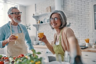 Carefree senior couple in aprons toasting each other with orange juice and preparing healthy dinner while spending time at home