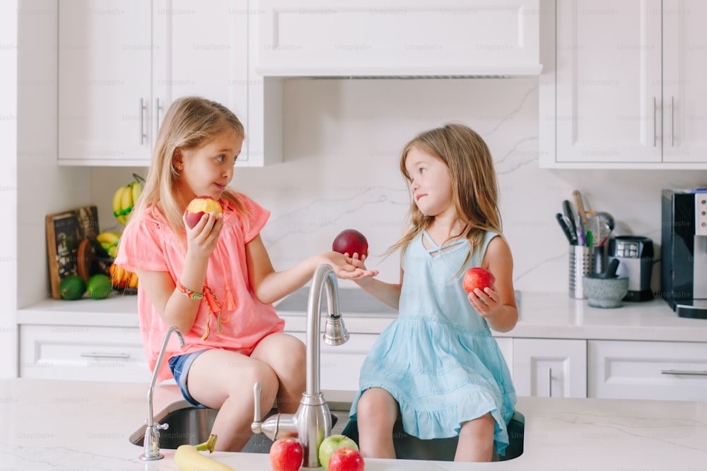 Caucasian children girls eating sharing fresh fruits sitting in kitchen sink. Happy family sisters siblings having snack. Organic food healthy delicious tasty meal for kids. Lifestyle authentic moment