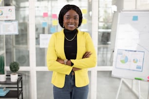 Front view of confident african woman in stylish clothing standing with crossed arms at office. Blur background of modern interior with flipchart and graphs.