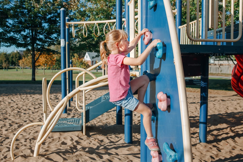Little preschool girl climbing rock wall at playground outside on summer day. Happy childhood lifestyle concept. Seasonal outdoor activity for kids. Strong girl female power.