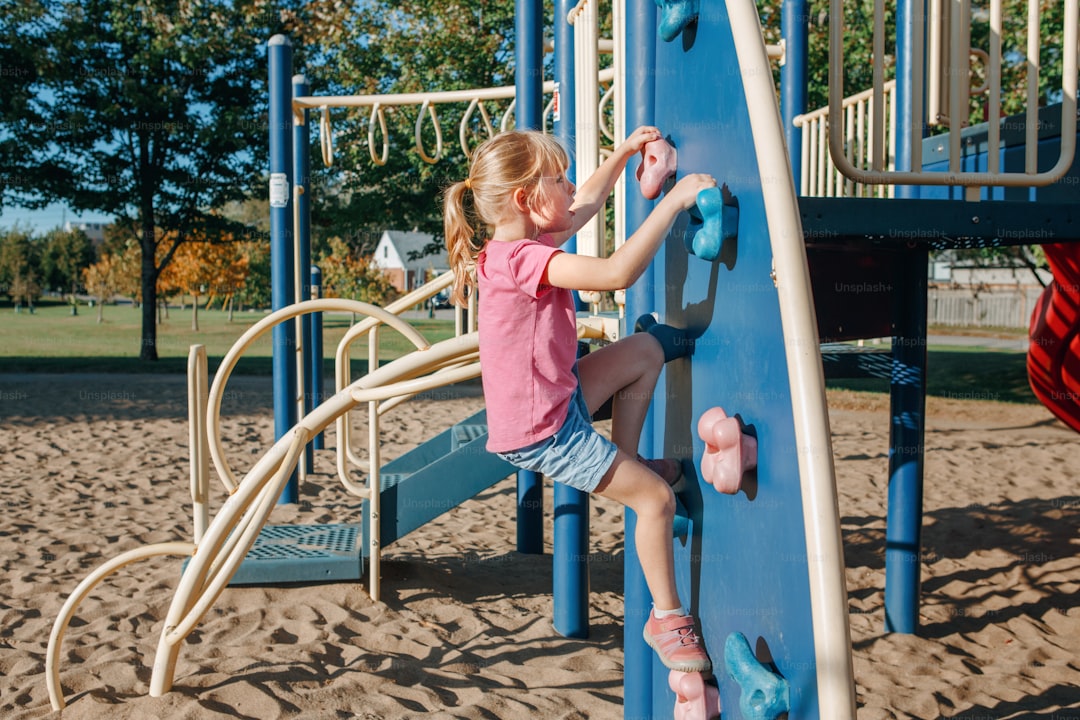 Little preschool girl climbing rock wall at playground outside on summer day. Happy childhood lifestyle concept. Seasona