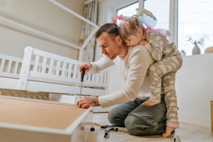小さな4歳の女の子は、父親が子供部屋のベッドの引き出しを組み立てたり修理したりするのを手伝います。