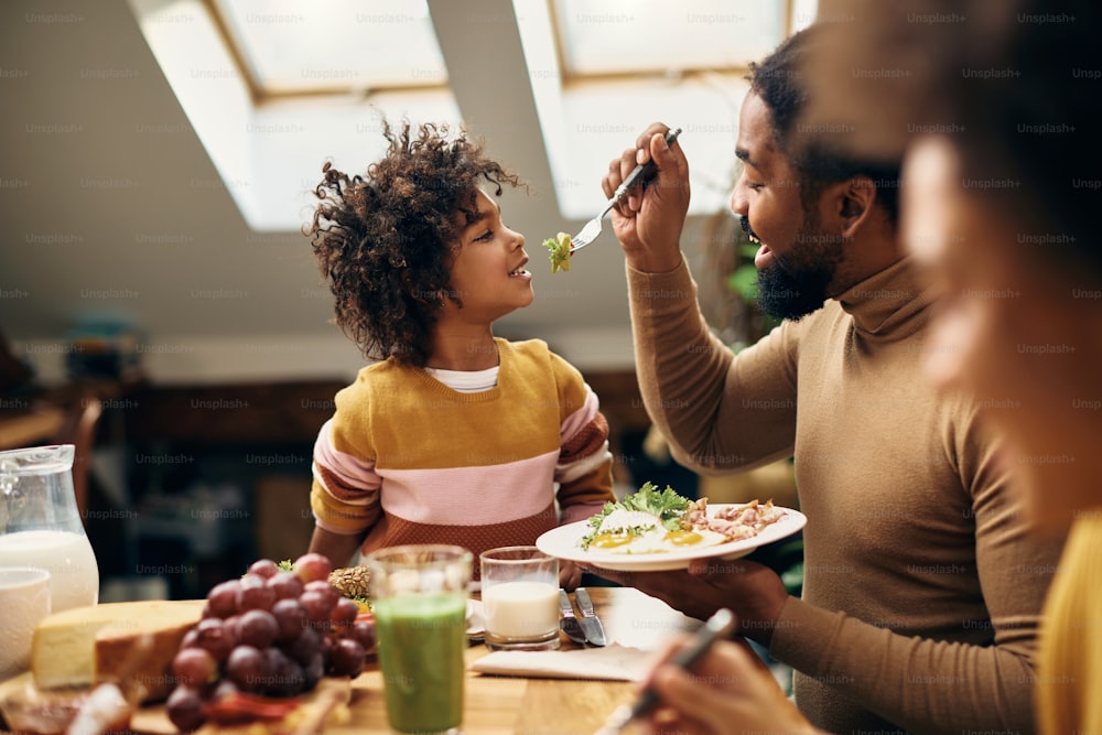 식탁에서 아버지에게 먹이를 주면서 즐거운 시간을 보내는 행복한 흑인 소녀.