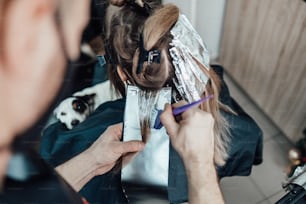 Hairdresser is dyeing female hair, making hair highlights to his client with a foil. They are wearing protective face mask as protection against virus pandemic.
