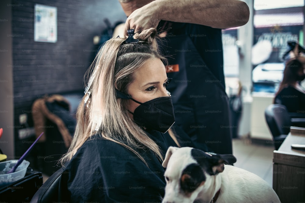 미용사는 여성의 머리카락을 염색하고 호일로 고객에게 머리카락 하이라이트를 만들고 있습니다. 그들은 바이러스 전염병으로부터 보호하기 위해 보호용 안면 마스크를 착용하고 있습니다.