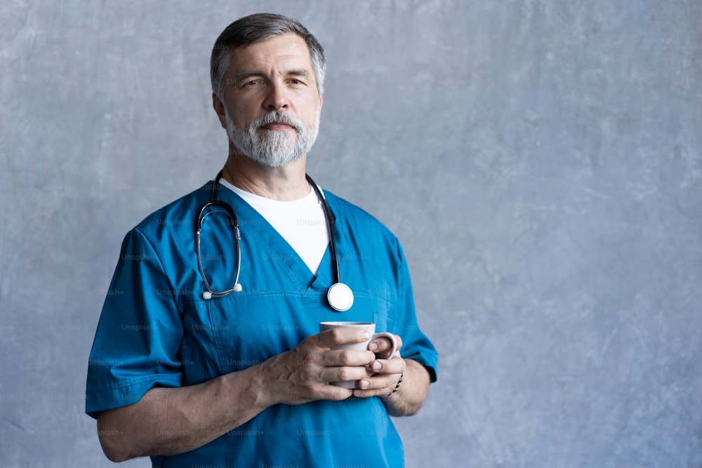 Ritratto di chirurgo maturo professionista che tiene la tazza, guardando la macchina fotografica mentre si trova contro lo sfondo grigio