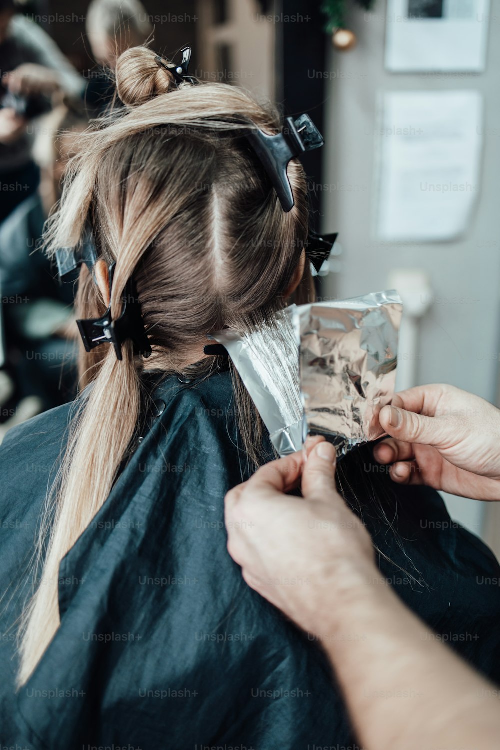 El peluquero tiñe el cabello femenino, haciendo reflejos para su cliente con una lámina. Llevan mascarilla protectora como protección contra la pandemia del virus.