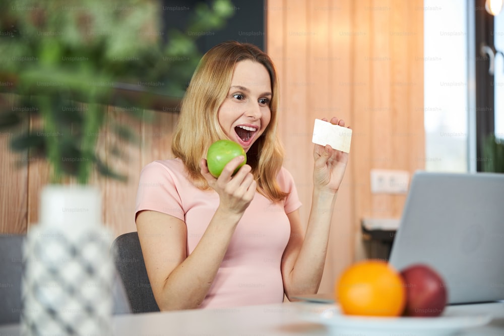 Cheerful lady sitting at the table with notebook and smiling while holding apple and credit card