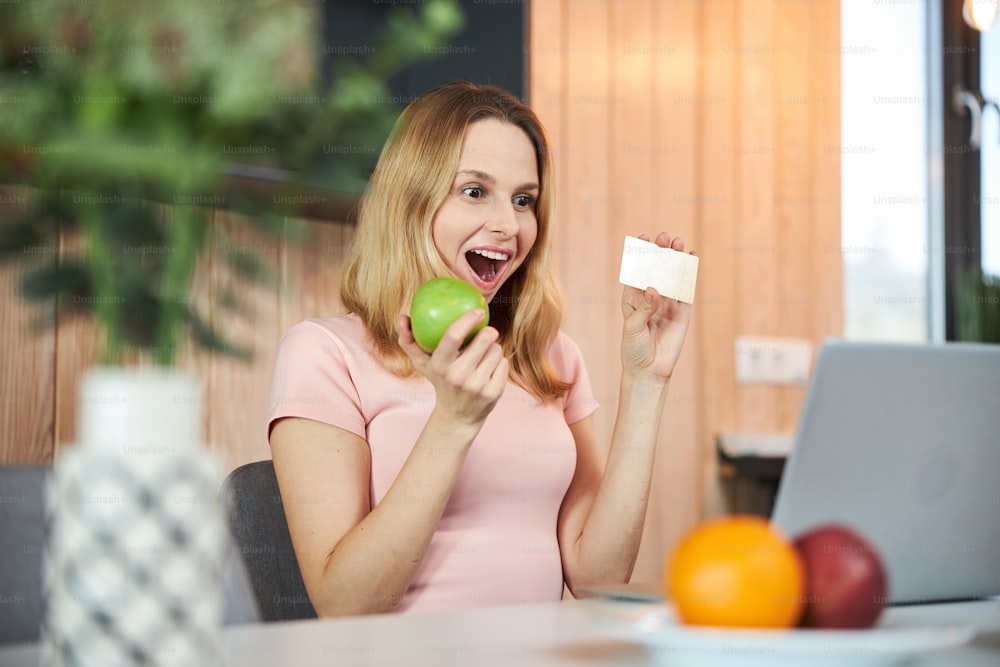 Señora alegre sentada en la mesa con cuaderno y sonriendo mientras sostiene la manzana y la tarjeta de crédito