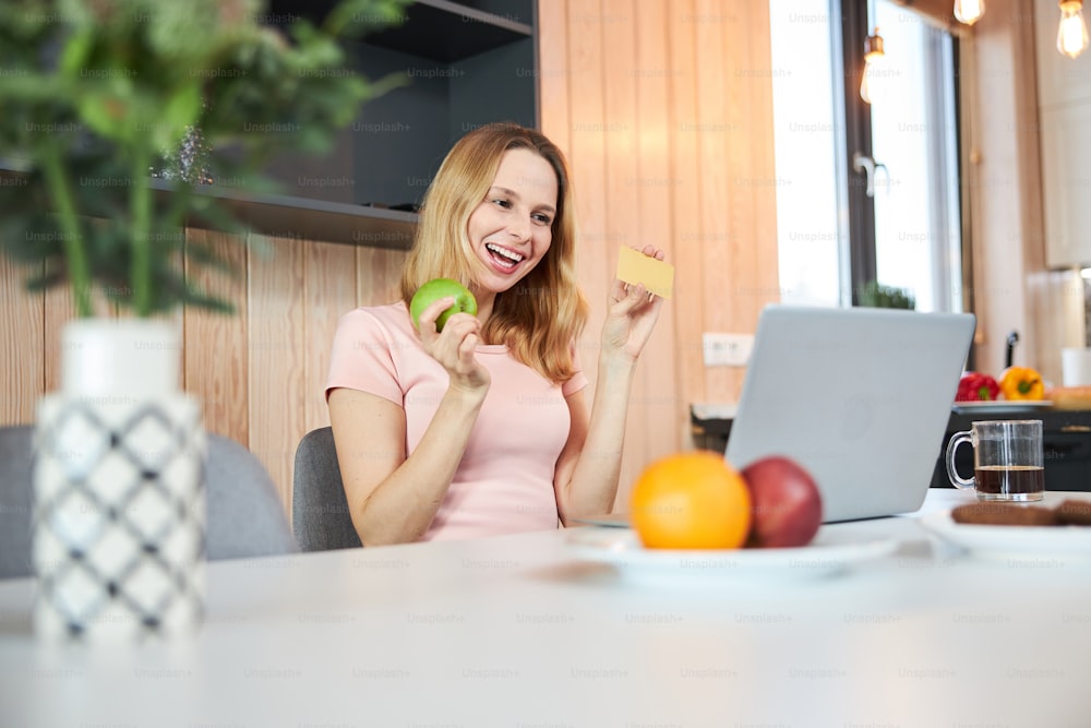 Dama encantadora sentada en la mesa con computadora portátil y sonriendo mientras sostiene la manzana y la tarjeta de crédito