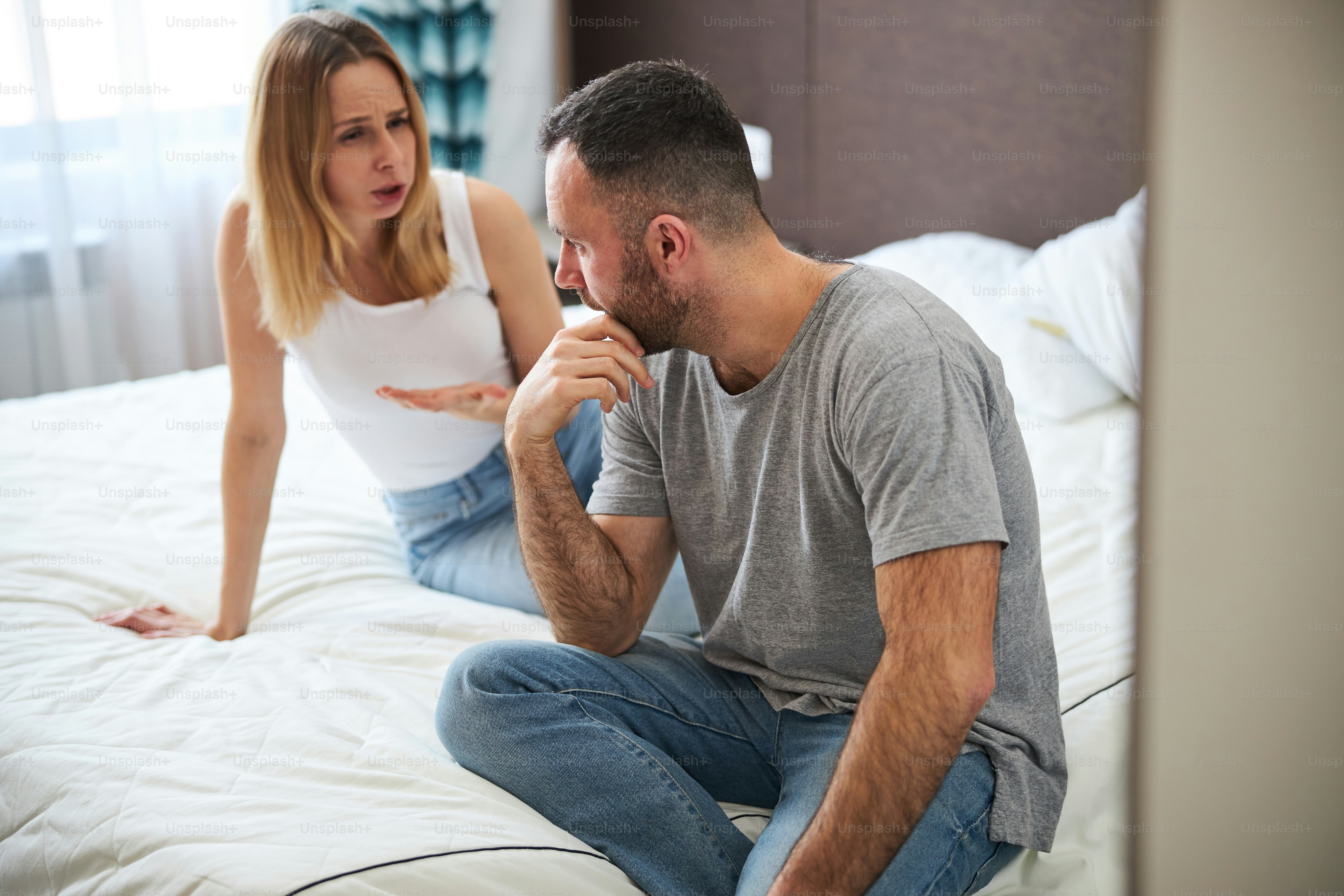 Displeased blonde lady sitting on bed and talking with husband while man touching his chin