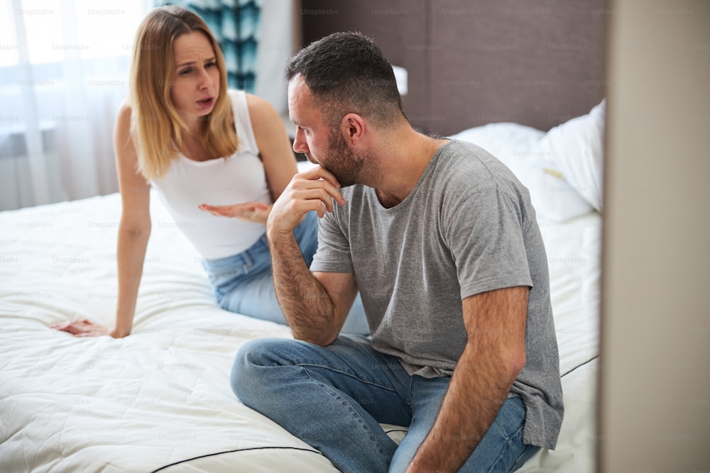 Displeased blonde lady sitting on bed and talking with husband while man touching his chin