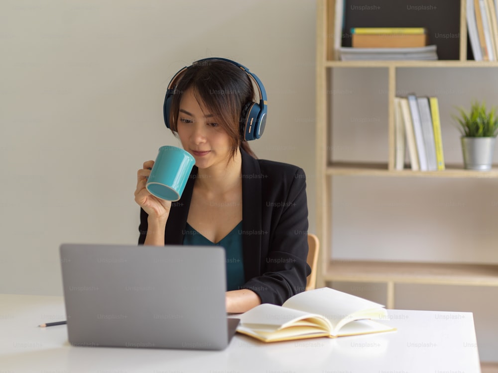 Retrato da empresária que trabalha em casa, usando laptop, fone de ouvido e bebendo café na sala do escritório em casa