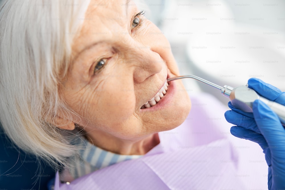 치과 전문가가 공기 물 스프레이 핸드피스로 치아를 청소하게 하는 은퇴한 여성의 클로즈업 사진