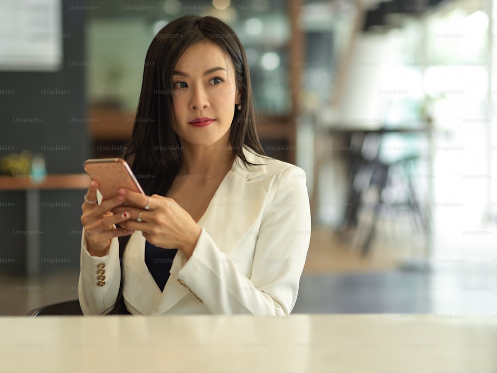 Ritratto di donna d'affari che tiene lo smartphone tra le mani e guarda in alto mentre è seduto nella stanza dell'ufficio