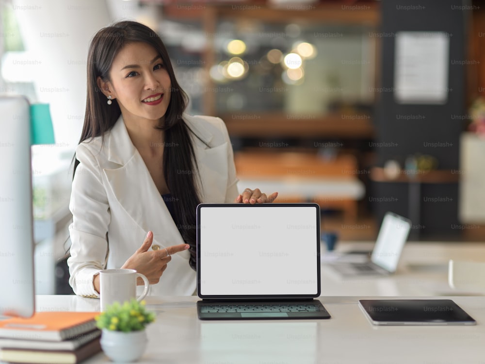 Nahaufnahme einer Geschäftsfrau in weißem Anzug, die ein digitales Tablet präsentiert und auf einen simulierten Bildschirm im Büroraum zeigt, Clipping-Pfad