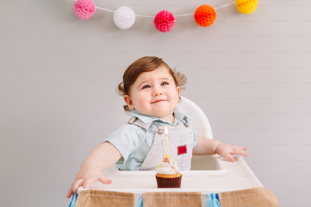 Felice sorridente carino bambino caucasico che festeggia il suo primo compleanno a casa. Bambino bambino bambino seduto in seggiolone mangiando gustoso dessert cupcake con candela accesa. Concetto di buon compleanno.