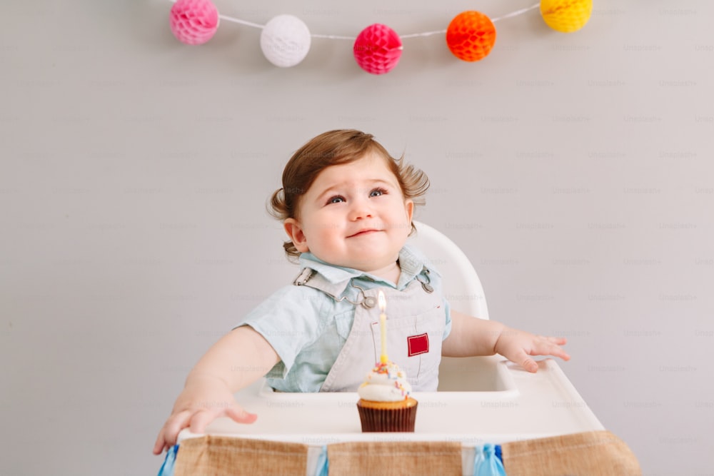 Glücklich lächelnder süßer kaukasischer Junge, der seinen ersten Geburtstag zu Hause feiert. Kind Kleinkind sitzt im Hochstuhl und isst leckeres Cupcake-Dessert mit brennender Kerze. Alles Gute zum Geburtstag.