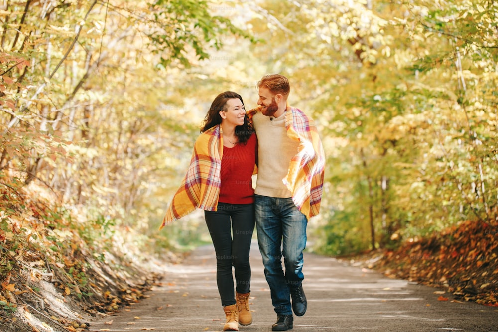 Beautiful couple man woman in love. Smiling laughing boyfriend and girlfriend wrapped in yellow blanket walking in park on autumn fall day. Togetherness and happiness. Authentic real people.