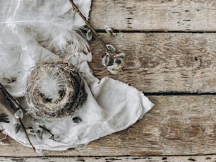 Rustic Easter flat lay with space for text. Bird nest on rustic background with feathers, pussy willow branches, cloth on aged wooden table. Stylish Easter rural still life, brown and grey colors