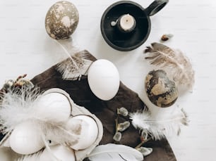 Natural easter eggs, feathers, pussy willow branches, candle on rustic cloth on white aged table. Easter flat lay. Stylish rural still life. Modern simple aesthetics, white and grey colors