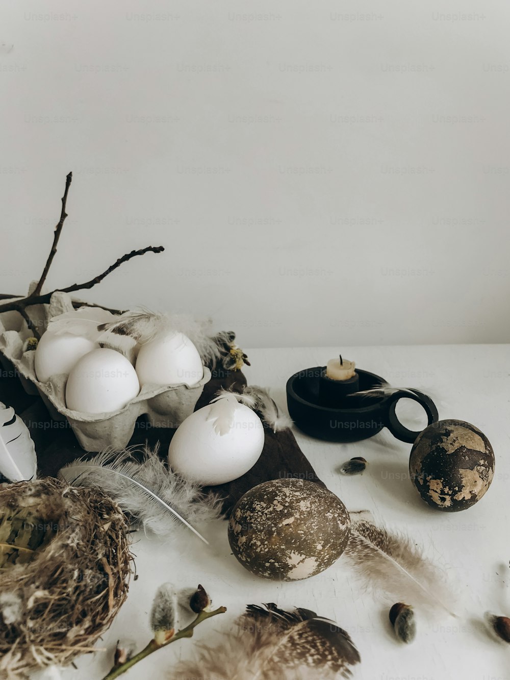Natürliche Ostereier, Federn, Weidenzweige, Nest und Kerze auf rustikalem Stoff auf weiß gealtertem Tisch. Stilvolles ländliches Osterstillleben. Moderne schlichte Ästhetik, weiße und graue Farben