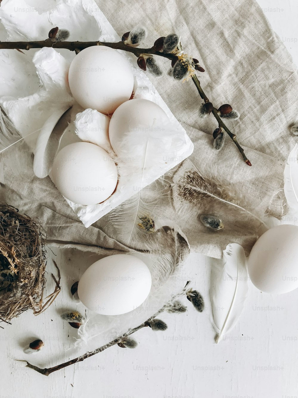 Natural easter eggs, soft feathers, pussy willow branches on rustic cloth on white aged table. Flat lay. Simple stylish rural Easter still life. Modern aesthetics, pastel beige and grey colors