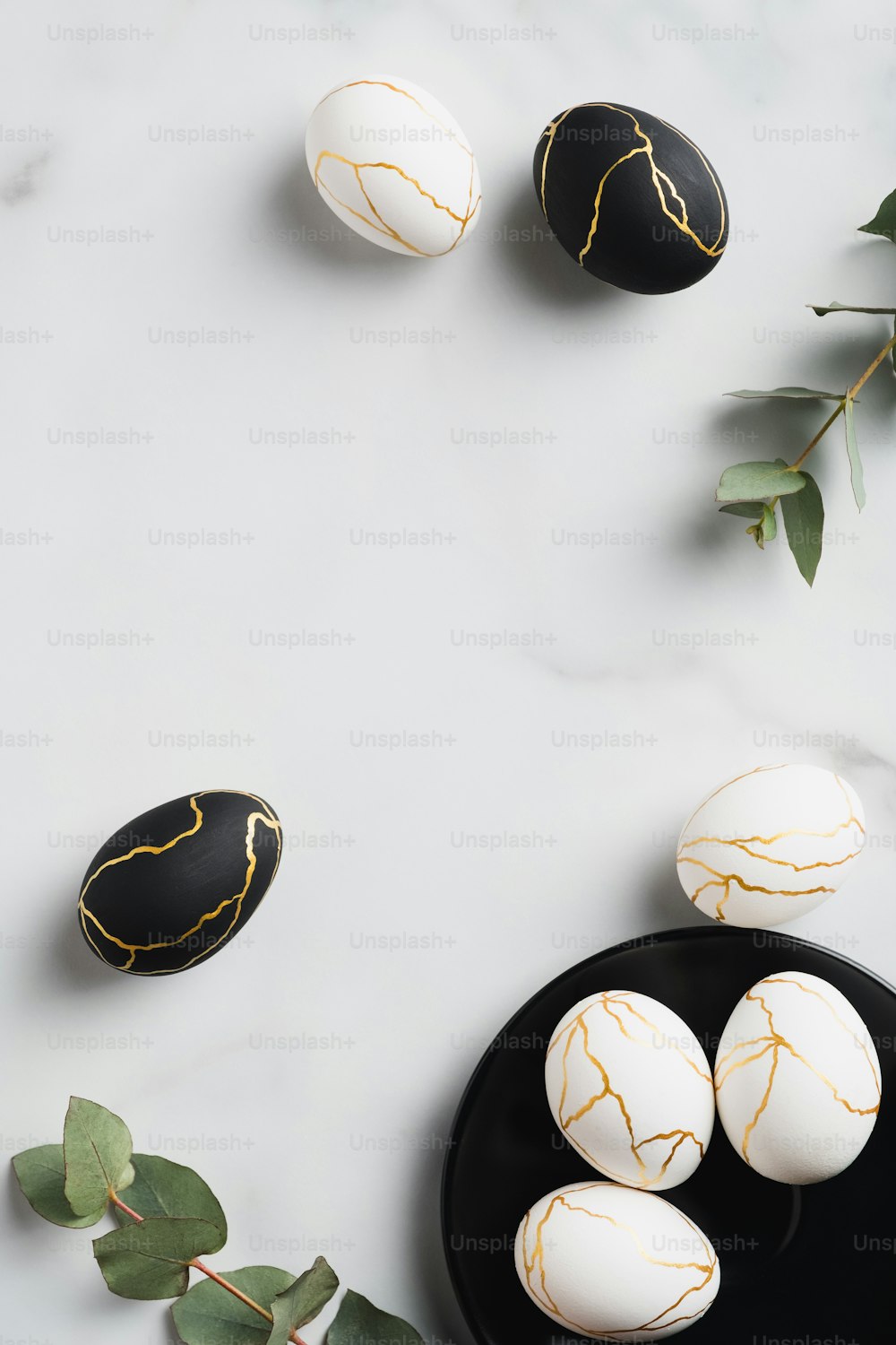 エレガントな白と黒のイースターエッグは、大理石のテーブルの上に金とユーカリの葉を飾っています。フラットレイ、上面図、垂直。