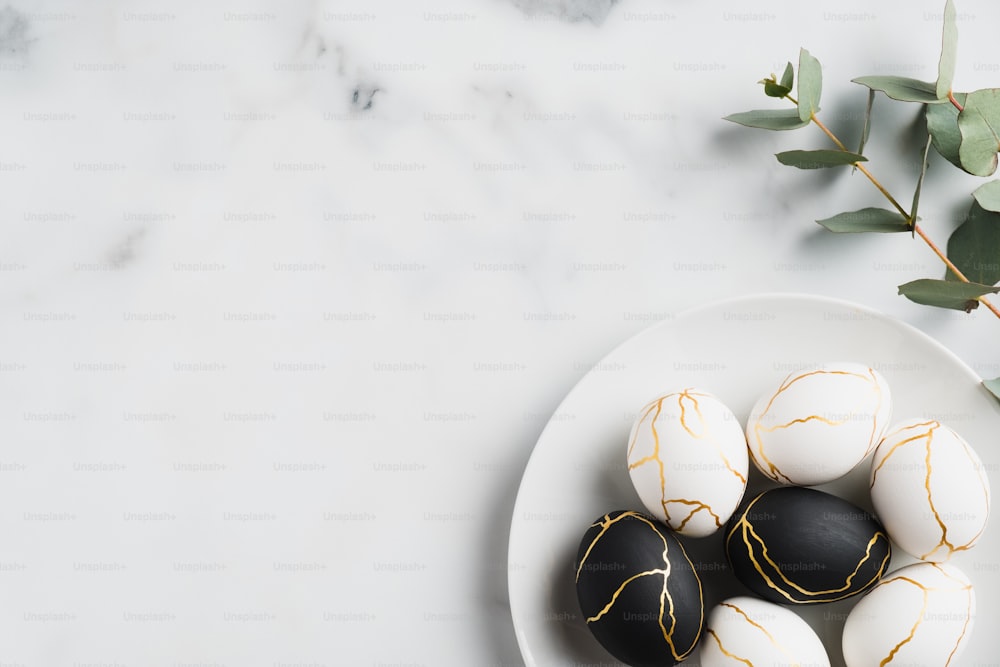 Luxury white and black Easter eggs decorated gold and eucalyptus leaves on marble table. Happy Easter card. Flat lay, top view, copy space.