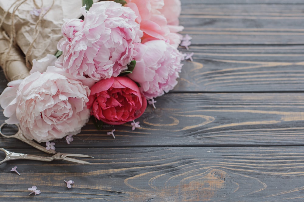 Beautiful stylish peony bouquet in paper, twine and scissors on rustic dark wooden background, copy space. Happy Mothers day greetings. Pink and white peonies flowers and lilac petals