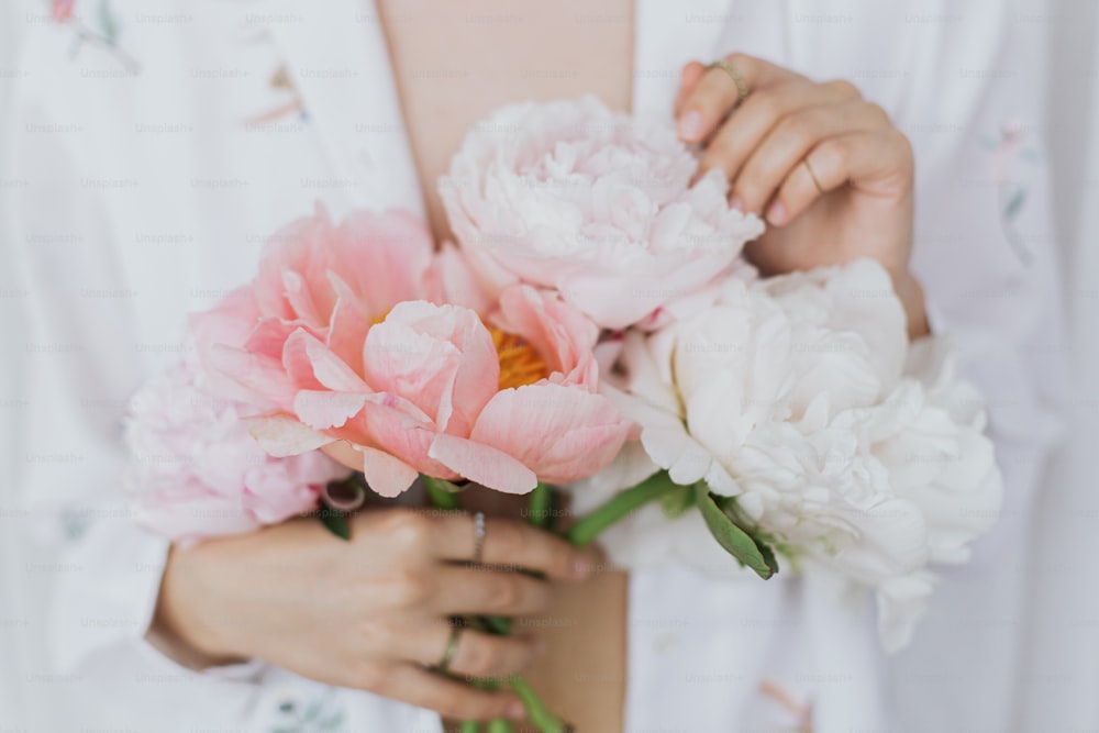 모란 꽃다발을 들고 아름 다운 세련 된 여자입니다. 보헤미안 꽃 셔츠를 입은 젊은 여성이 분홍색과 흰색 모란 꽃을 손에 부드럽게 들고 있다. 관능적 인 부드러운 이미지. 봄의 미학