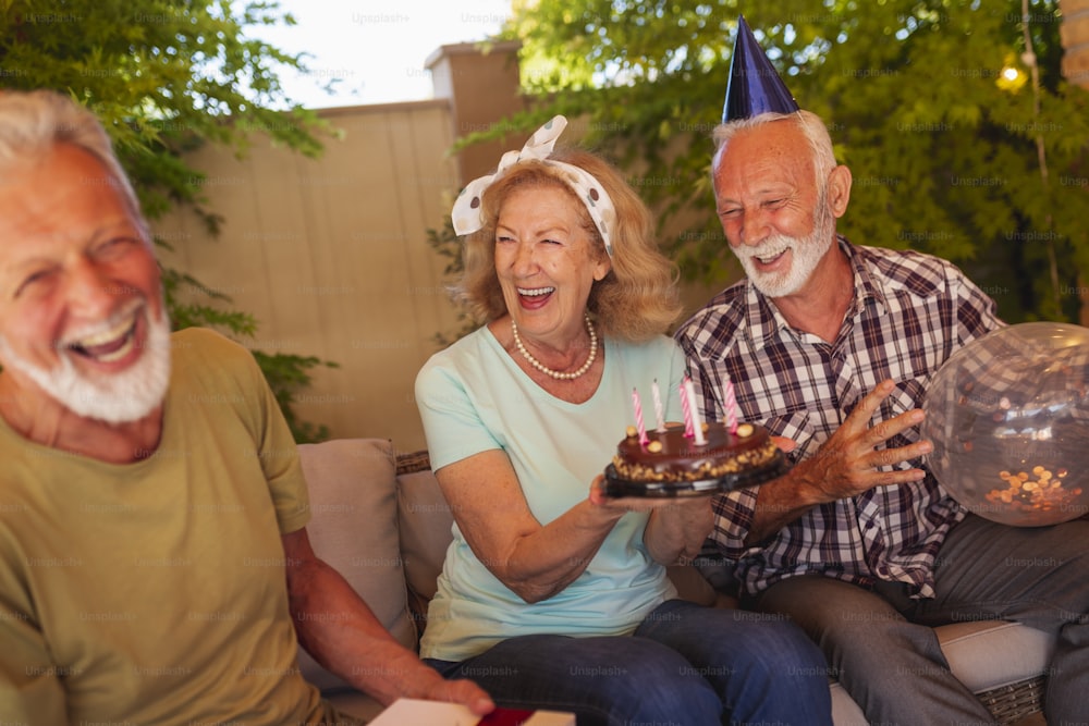Grupo de alegres amigos mayores que se divierten en una fiesta de cumpleaños, anfitrión de la fiesta que sostiene un pastel de cumpleaños después de pedir un deseo y soplar las velas