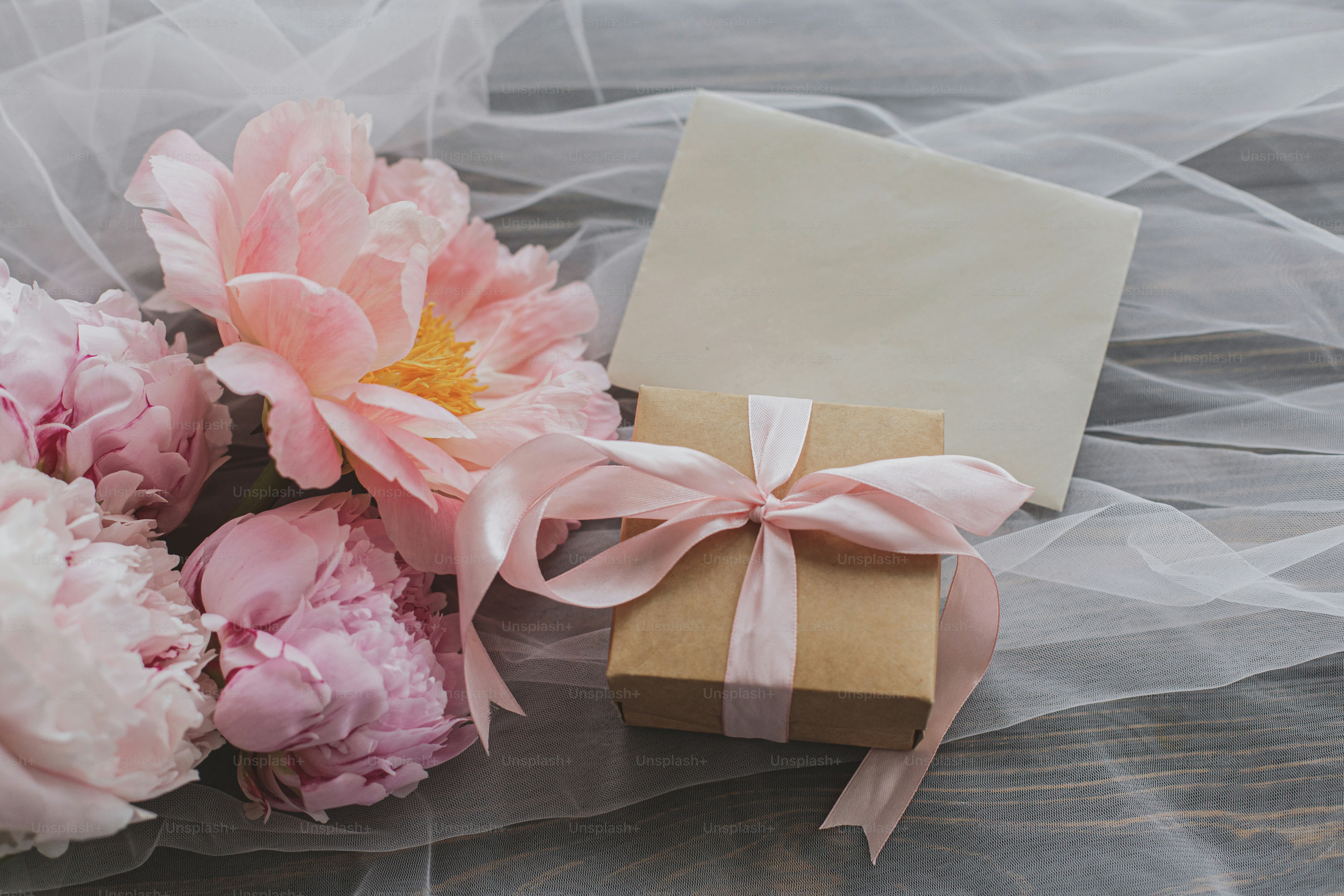 Beautiful peony bouquet, gift box and greeting card on soft tulle fabric on dark wooden background, top view. Pink and white peonies flowers and stylish present. Happy Mothers day