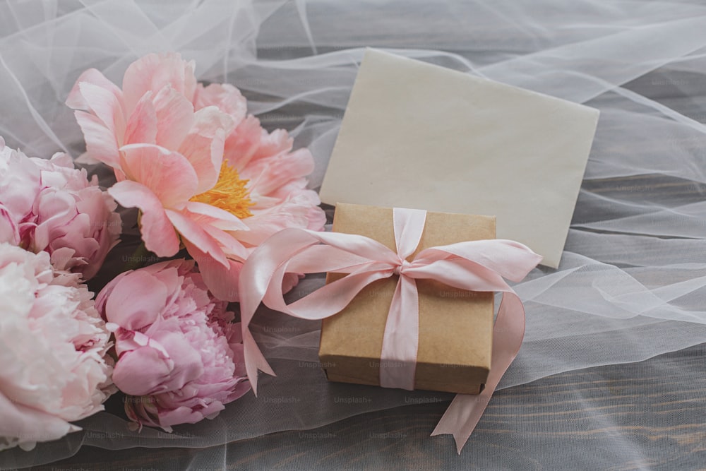 Beautiful peony bouquet, gift box and greeting card on soft tulle fabric on dark wooden background, top view. Pink and white peonies flowers and stylish present. Happy Mothers day