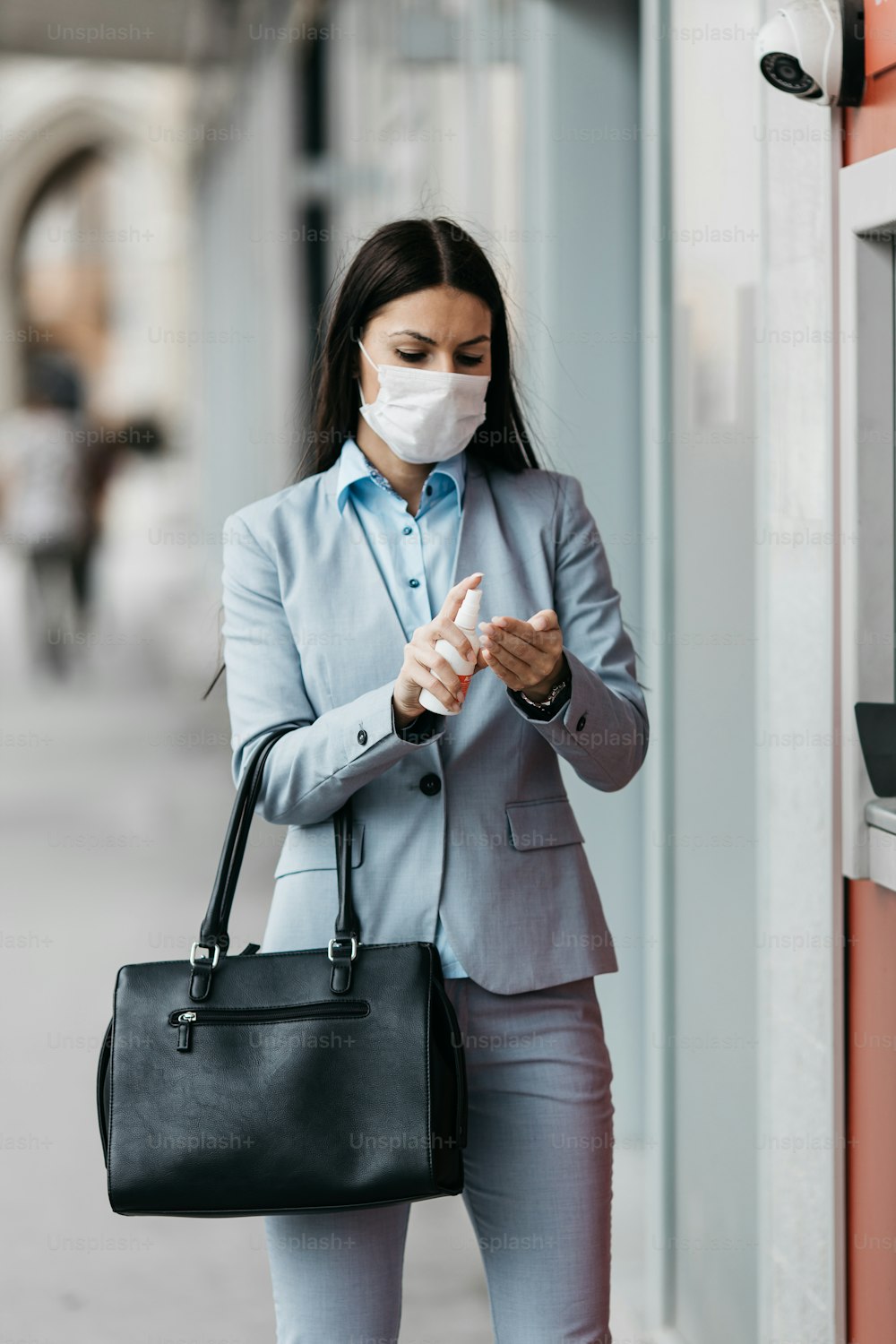 Elegante Geschäftsfrau mit Schutzmaske, die auf der Stadtstraße steht und am Geldautomaten Bargeld abhebt. Corona- oder Covid-19-Virus-Pandemie-Konzept.